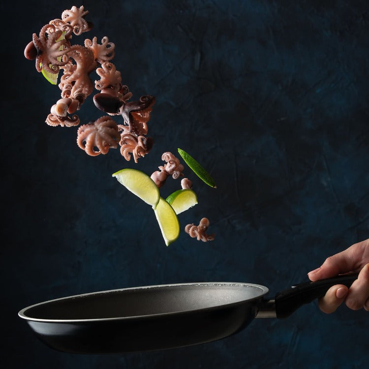 Dynamic image of baby octopus being flipped in a frying pan, accompanied by green lime slices. A visually engaging representation of fresh and ready-to-cook seafood