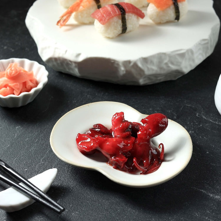 Small plate of seasoned baby octopus (Chuka Idako) on a dark background, served with sushi and pickled ginger in the background