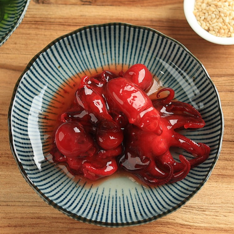 Bowl of seasoned baby octopus (Chuka Idako) with a glossy red marinade, placed on a wooden table with sesame seeds in the background