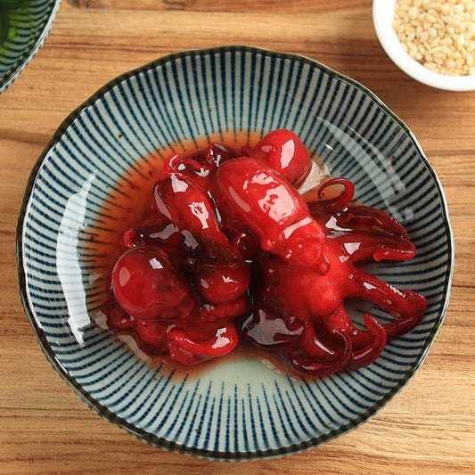 Bowl of seasoned baby octopus (Chuka Idako) with a glossy red marinade, placed on a wooden table with sesame seeds in the background