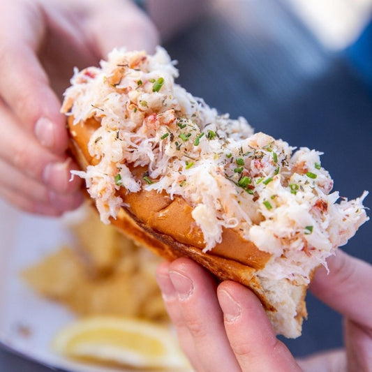 A close-up of a hand holding a crab roll sandwich filled generously with shredded imitation crab meat mixed with herbs and a light dressing, served in a toasted bun. The sandwich is a casual yet delicious way to enjoy seafood flavors