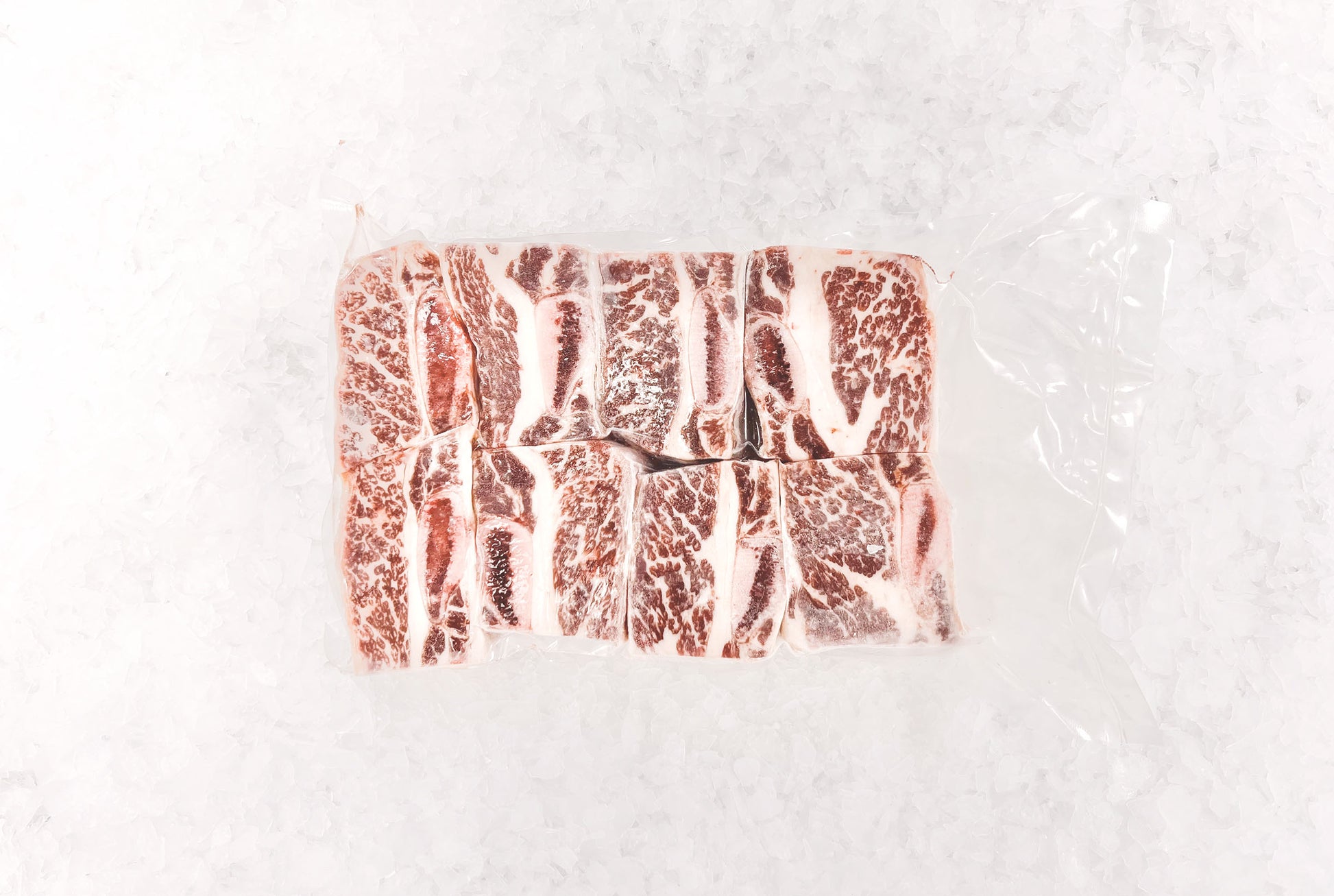 This image features a clear plastic-wrapped package of beef short rib cuts, neatly arranged on crushed ice. The meat displays a rich marbling of fat, emphasizing its quality and texture