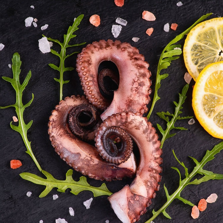 Freshly cut raw tako legs arranged artistically on a black slate, garnished with lemon slices and rock salt