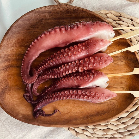 Skewered tako legs prepared for grilling, displayed on a wooden serving board, emphasizing their natural pink and purple hues