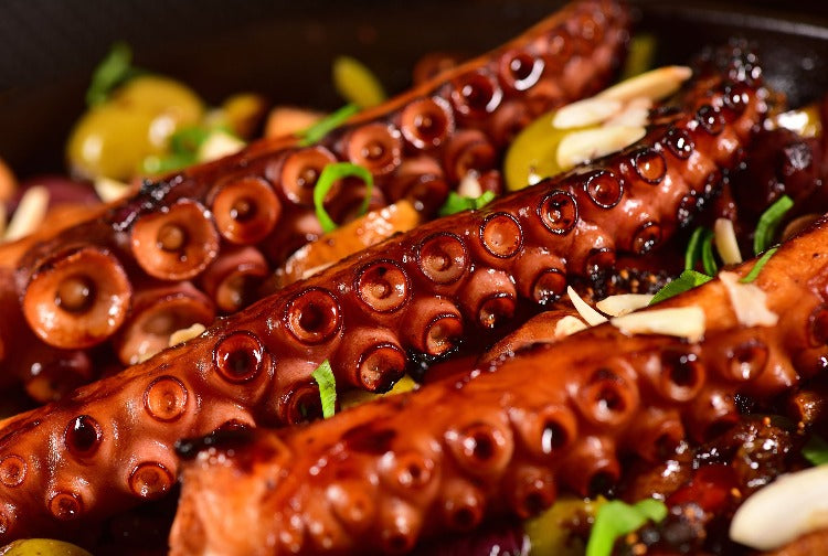 Close-up of grilled tako legs, beautifully charred and garnished with green herbs and garlic, served on a rustic pan