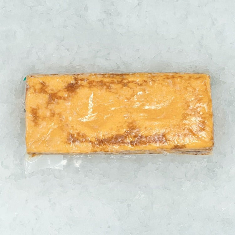 An overhead view of a block of tamagoyaki, still in its plastic wrap, which shows the fine layers and fluffy texture of the omelette, set against a background of crushed ice that enhances the visual appeal of the product being fresh and ready for consumption