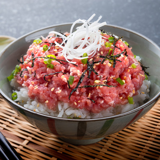A bowl of yellowfin tuna over rice, commonly known as tuna donburi or "tekka don". The ground tuna is sprinkled with sesame seeds, finely chopped seaweed, and green onions, served over steamed white rice, garnished with a side of radish sprouts. This dish highlights the versatile use of ground tuna in Japanese cuisine