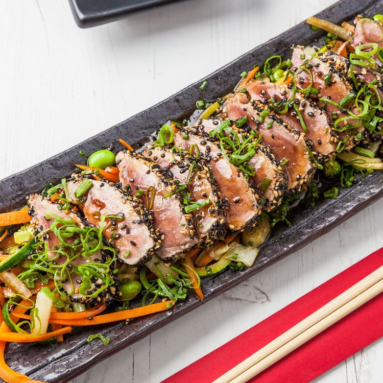 Sliced Katsuo tataki (grilled skipjack tuna) served on a black plate with fresh vegetables. The tuna is lightly seared, topped with sesame seeds and green onions, showcasing a vibrant and appetizing presentation