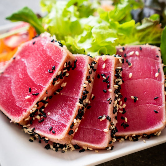 Close-up view of seared Katsuo tataki (skipjack tuna) slices, coated with black and white sesame seeds, served with a fresh vegetable salad. The tuna is tender and pink in the center, emphasizing its freshness and quality