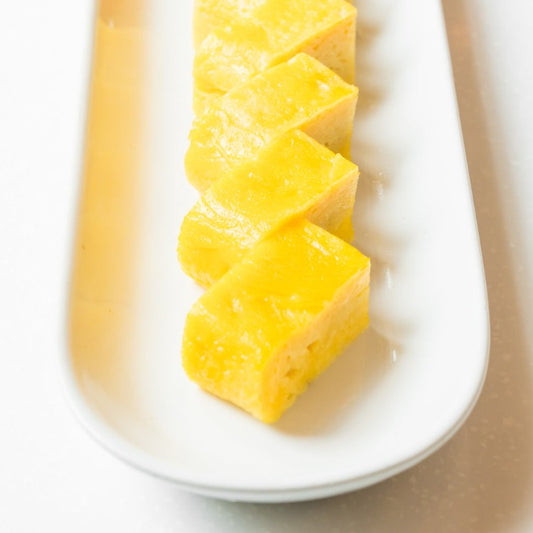 Slices of tamagoyaki arranged neatly on a white ceramic plate, each piece showing the layered and airy structure typical of this Japanese dish. The bright yellow color of the omelette makes it stand out, ideal for a visually appealing presentation at any meal