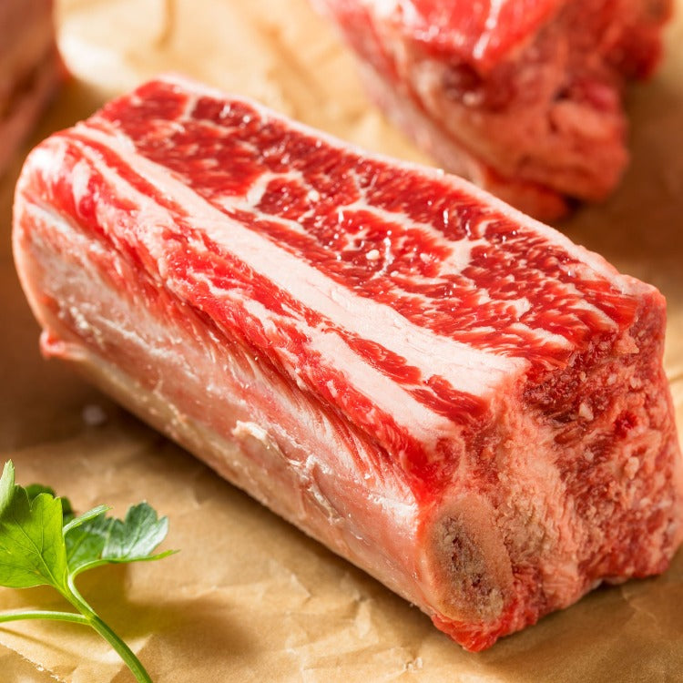 Another close-up image of raw beef short rib, similarly placed on butcher paper. The meat showcases deep red color and extensive marbling, ready for cooking or grilling, with a fresh parsley leaf adding a touch of green