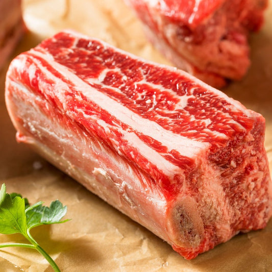 Another close-up image of raw beef short rib, similarly placed on butcher paper. The meat showcases deep red color and extensive marbling, ready for cooking or grilling, with a fresh parsley leaf adding a touch of green