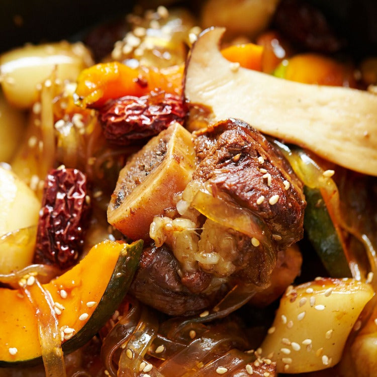 This is a close-up image of a Korean dish, Galbi Jjim, featuring braised beef short ribs with vegetables and dried red chili peppers. The meat is tender and glazed with a shiny, savory sauce, garnished with sesame seeds