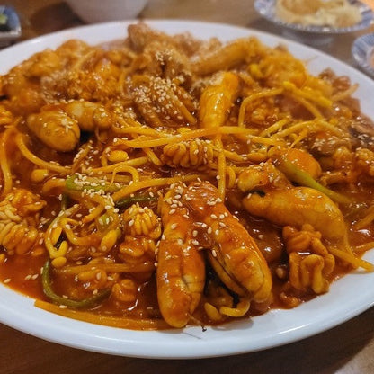 This image shows a hearty Korean seafood dish featuring pollock roe, shrimp, and squid coated in a spicy, vibrant red sauce. The dish is garnished with sesame seeds and thinly sliced vegetables, served on a white plate