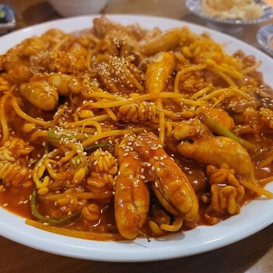 This image shows a hearty Korean seafood dish featuring pollock roe, shrimp, and squid coated in a spicy, vibrant red sauce. The dish is garnished with sesame seeds and thinly sliced vegetables, served on a white plate