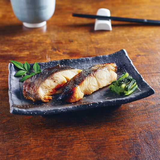 Exquisitely grilled Black Cod Kasuzuke served on a dark ceramic plate, accompanied by steamed greens, showcasing its glossy, caramelized surface.