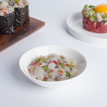 A bowl of seasoned chopped octopus mixed with vegetables, served as Tako Wasabi. The dish includes green peppers, red bell peppers, and sesame seeds, placed on a white background