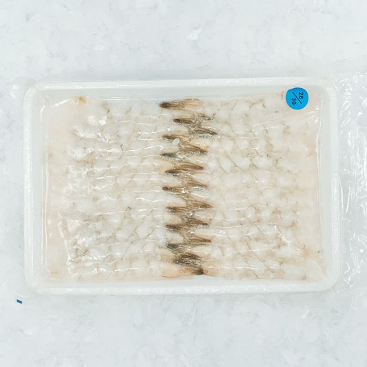 This image shows a tray of nobashi ebi (stretched shrimp), neatly lined up and presented on a white styrofoam tray, covered with a clear plastic wrap. The shrimp are uncooked, showcasing their translucent and pale pink hues. The tray is labeled with a blue circular sticker indicating the product number or price. The background features crushed ice, emphasizing the freshness and chilled storage of the seafood