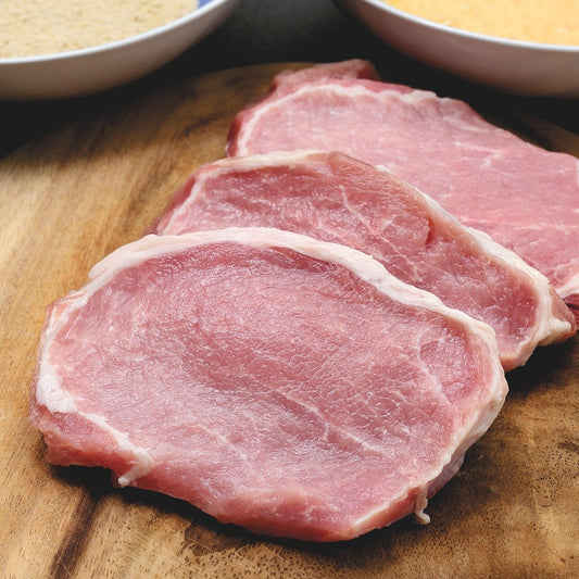 Raw pork steaks on a wooden cutting board with bowls of flour and breadcrumbs in the background.
